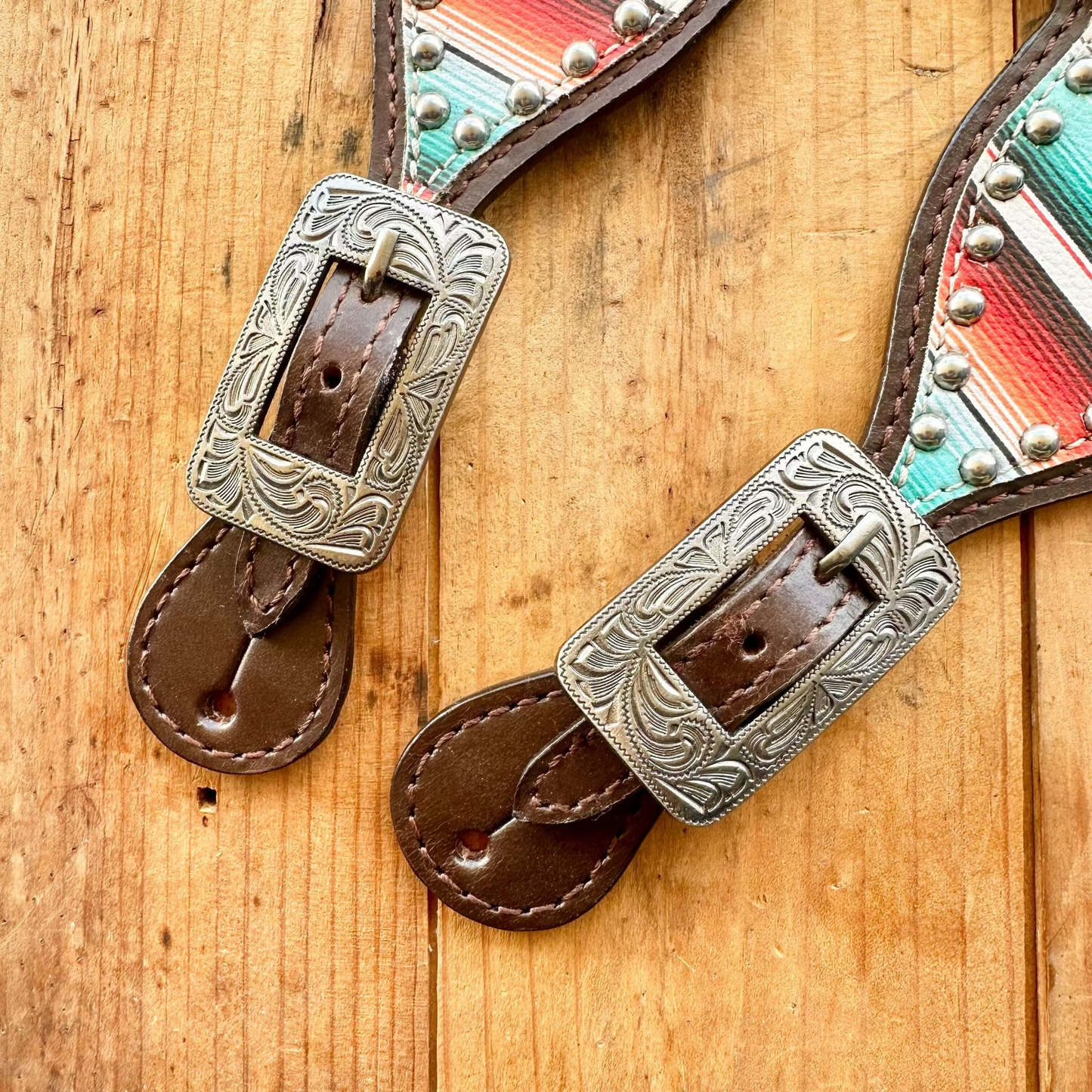 Two western-style leather straps laid on a wooden surface, each with an engraved silver-tone buckle, brown leather ends, and a multicolor striped fabric section with silver studs.