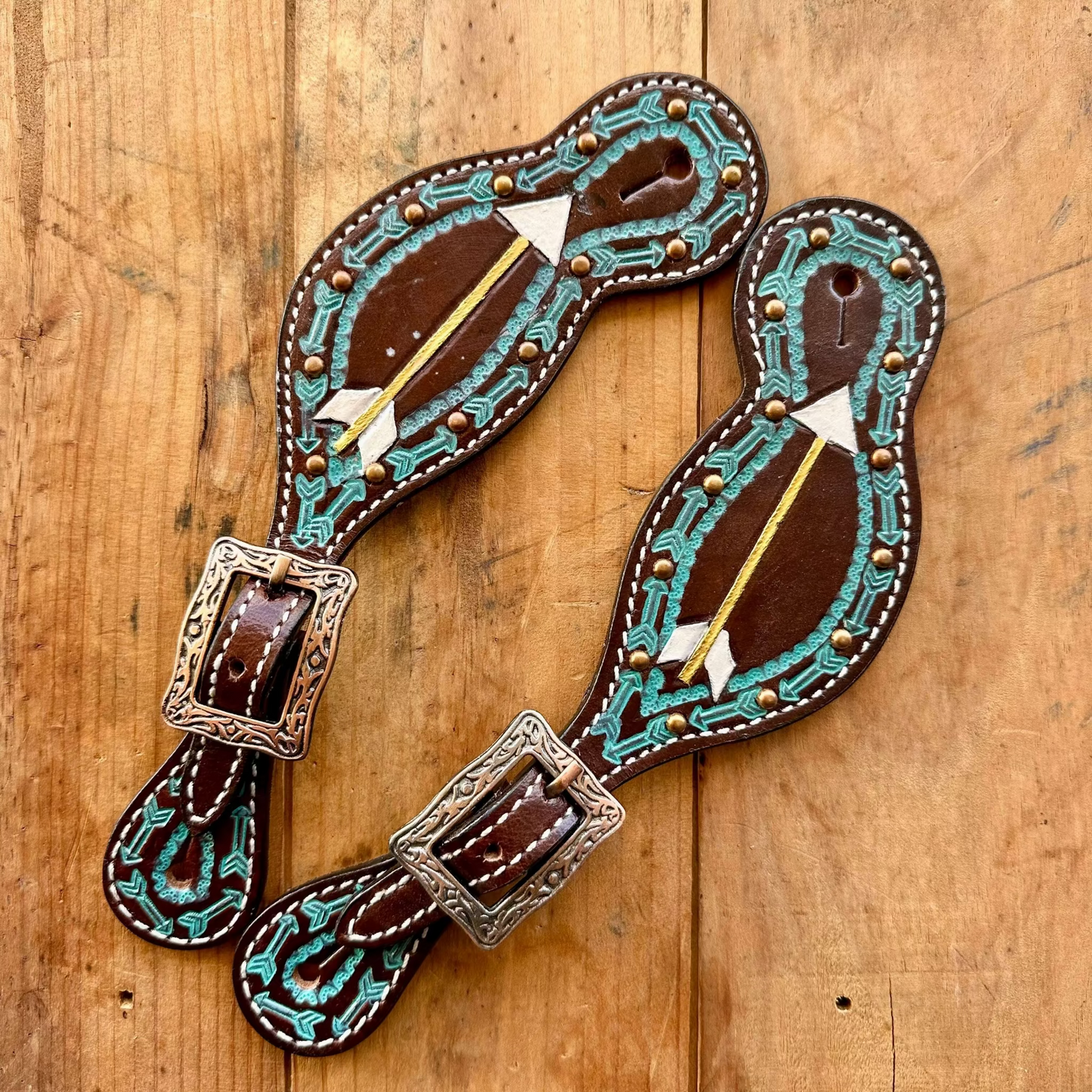 A pair of hand-tooled leather western boot pull tabs with turquoise tassel beadwork, white arrow inlays, and silver-tone buckles, displayed on a rustic wooden background.