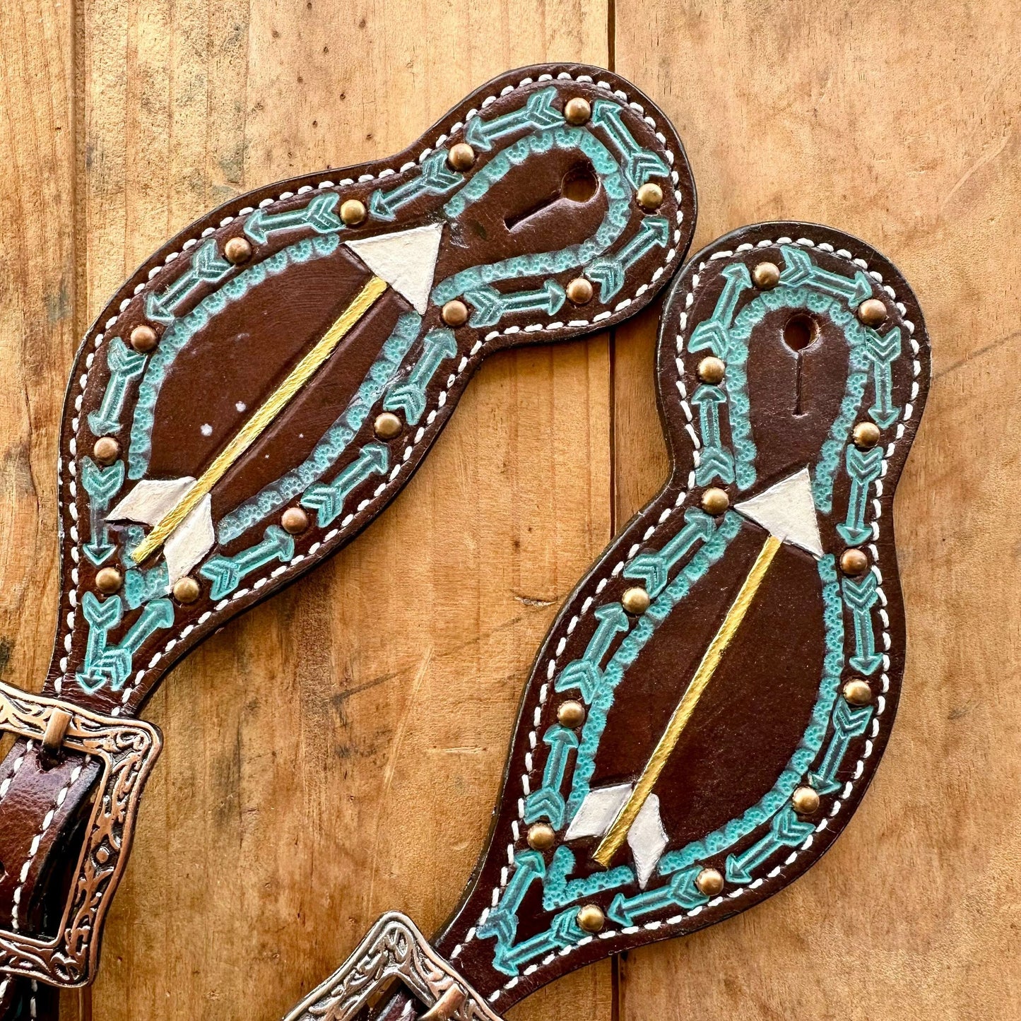 A pair of handcrafted brown leather western-style straps with intricate turquoise and white geometric beadwork, gold-toned arrow inlays, and brass studs, displayed on a rustic wooden background.