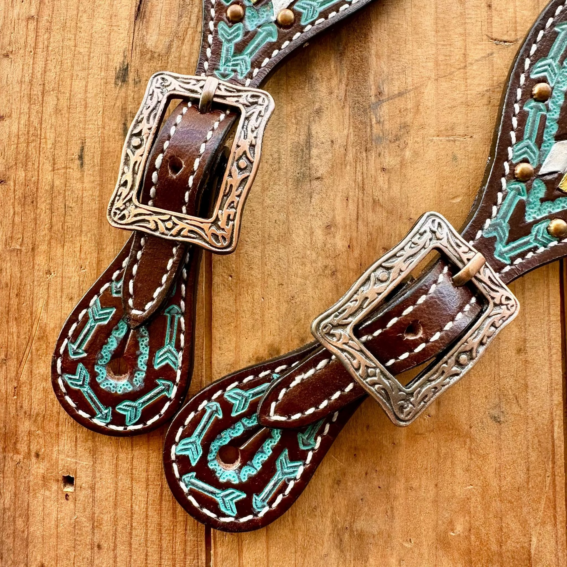 A pair of brown leather western-style belts with turquoise and white embroidered detailing and engraved silver-tone buckles, displayed on a rustic wooden surface.