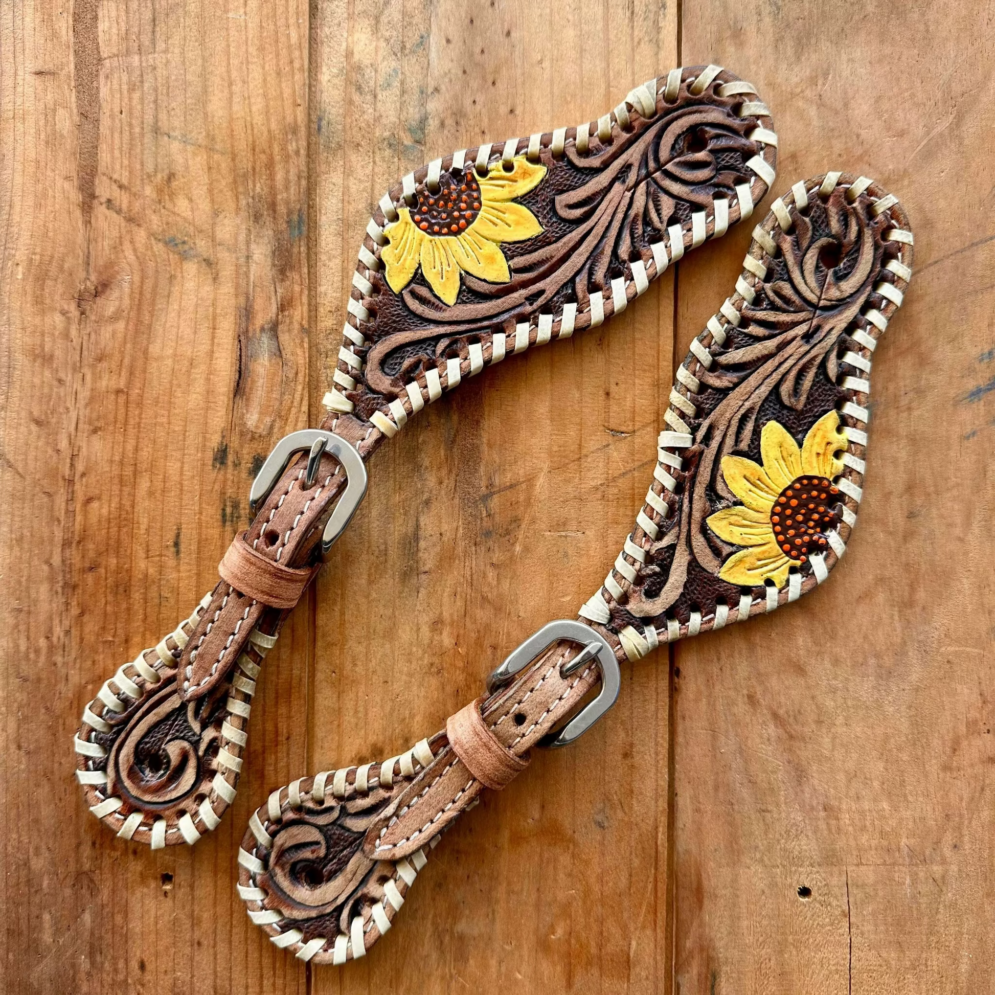 Two hand-tooled leather strap accessories with floral sunflower designs and braided trim, displayed on a rustic wooden surface.