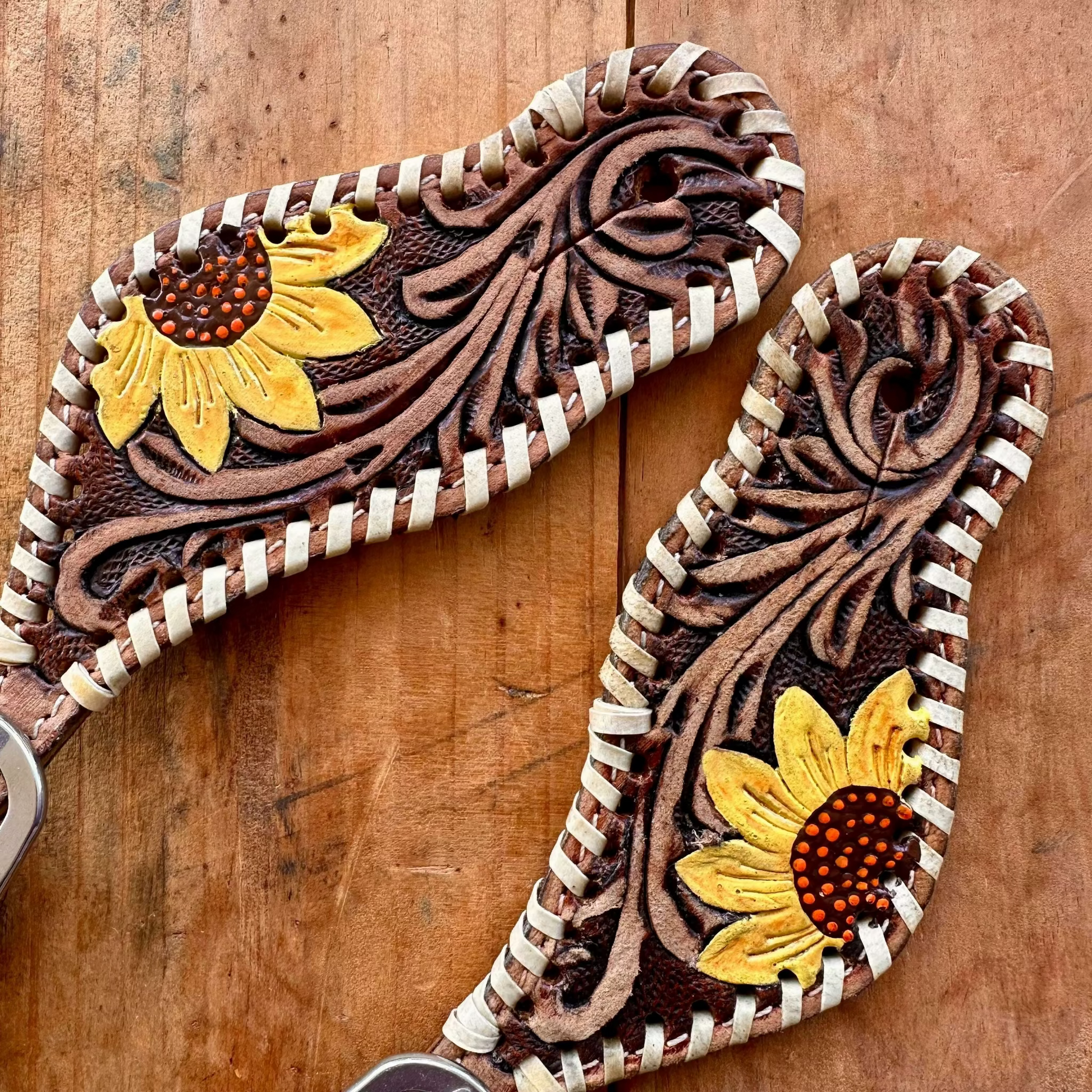 Two handcrafted tooled leather strap charms with carved floral designs and yellow sunflower accents, displayed on a rustic wooden surface.