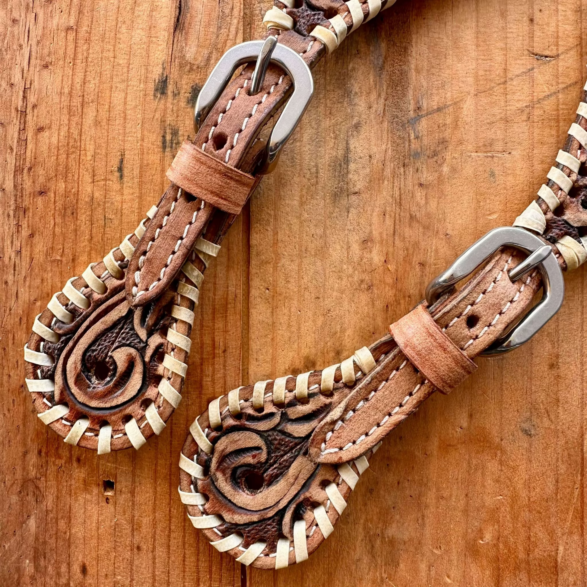 A close-up of a leather strap or belt with tooled floral detailing, braided cream stitching, and silver metal buckles, laid on a rustic wooden surface.