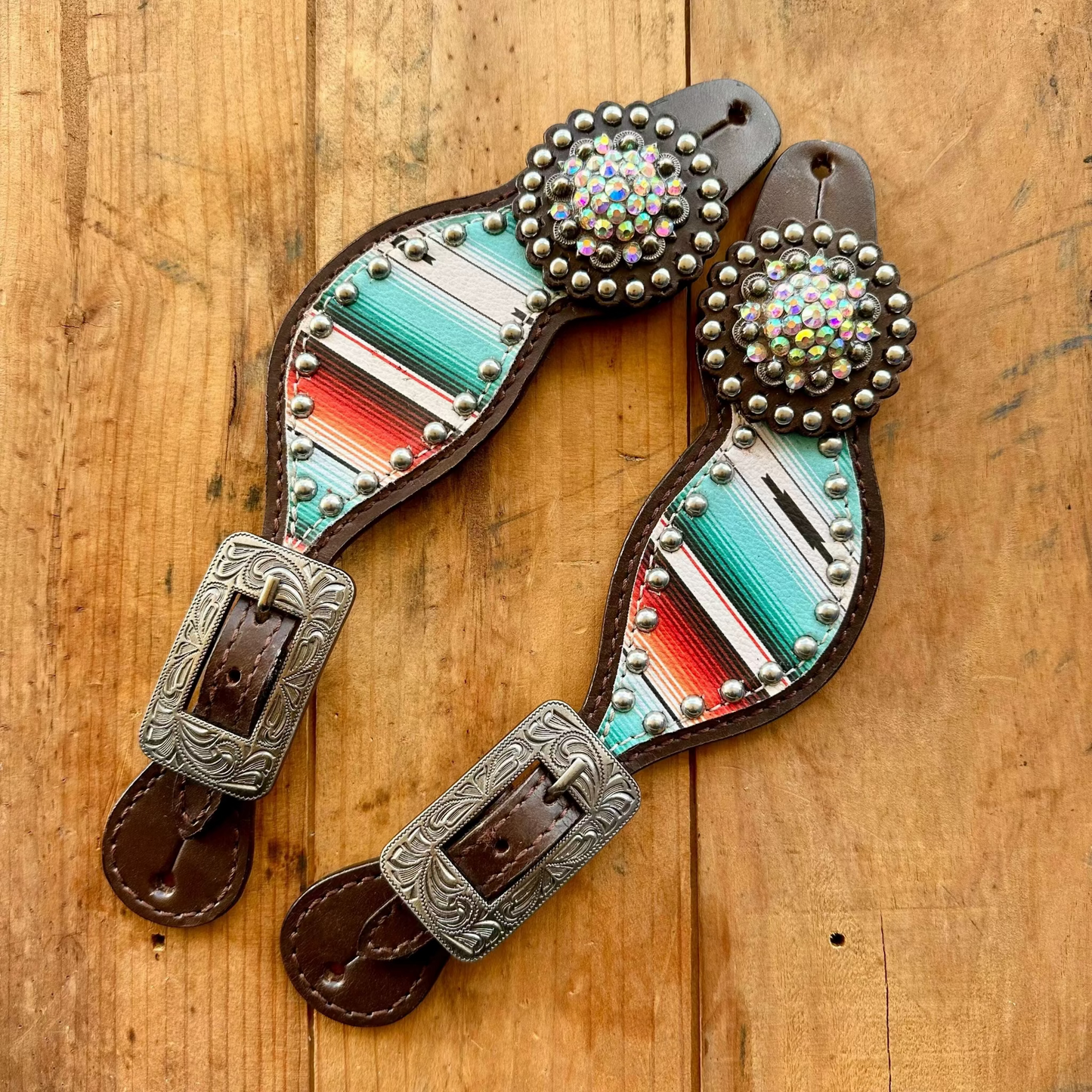 A pair of western-style leather strap accessories with silver-tone buckles, silver studs, and turquoise, red, white, and blue striped inlays, displayed on a rustic wooden surface.