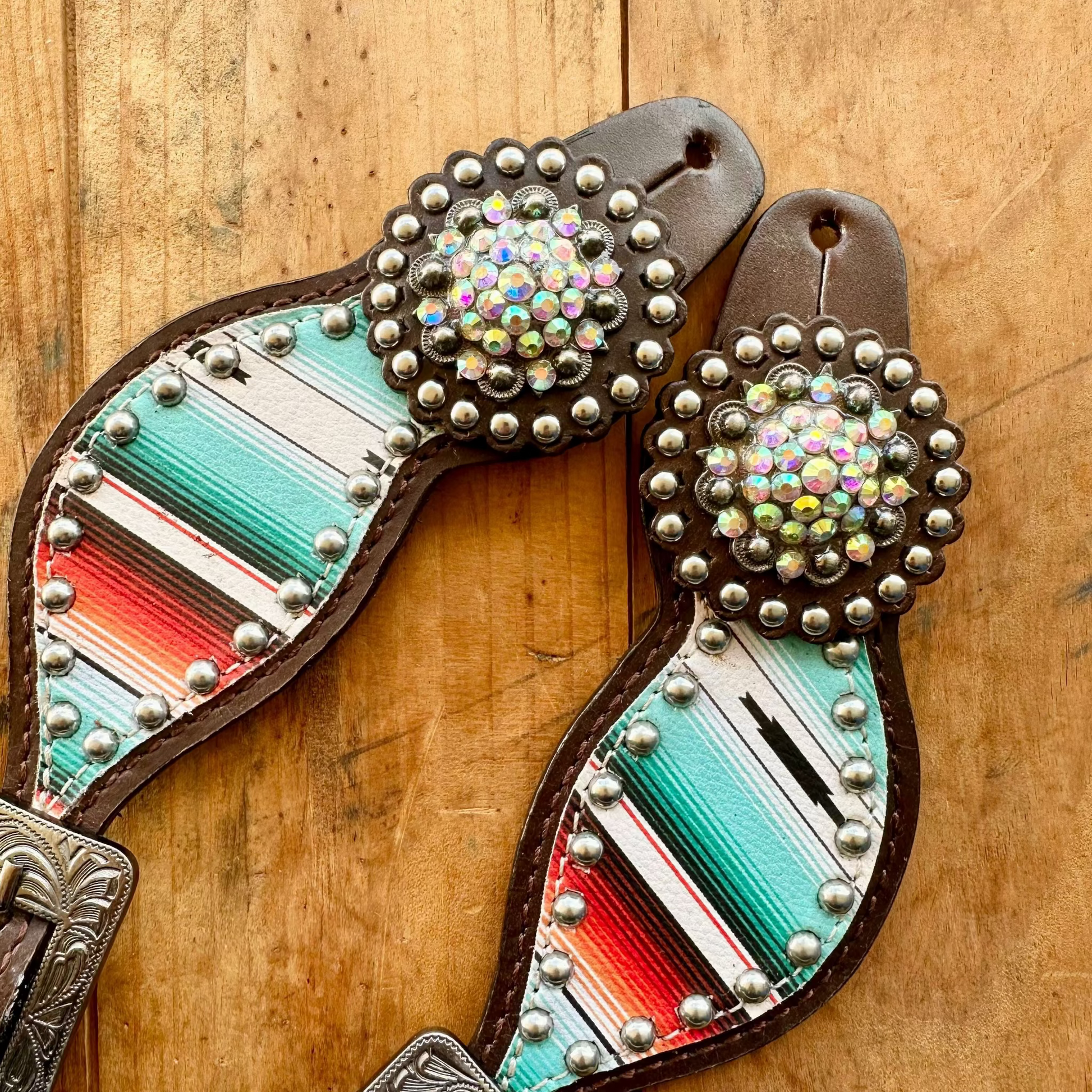 A pair of western-style leather strap accessories with turquoise, red, white, and brown striped fabric panels, silver studs, and iridescent rhinestone conchos, displayed on a wooden surface.