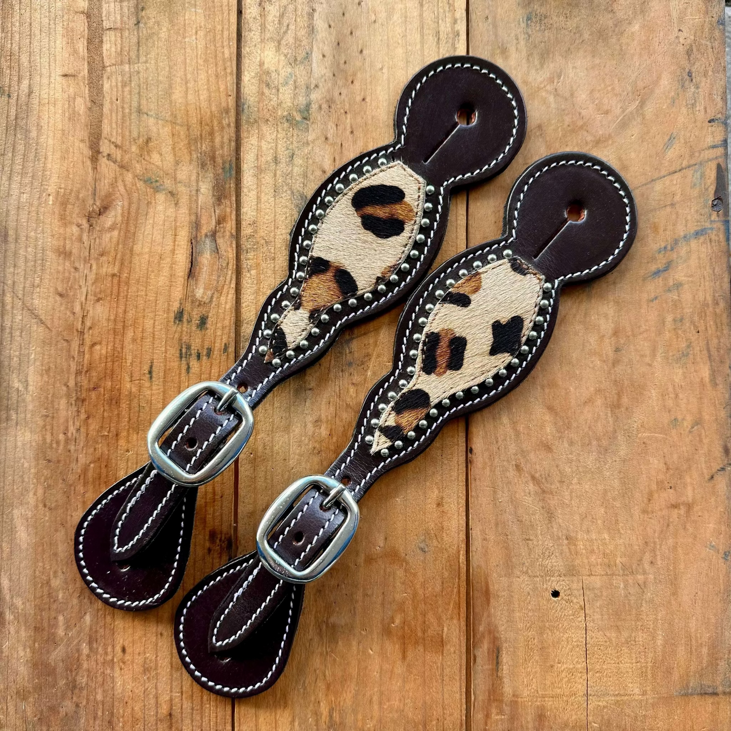 Two leather strap accessories with dark brown edges, white stitching, and a central tan-and-black leopard print panel, each ending in a dark brown loop and secured by silver-tone metal buckles, displayed on a rustic wooden surface.