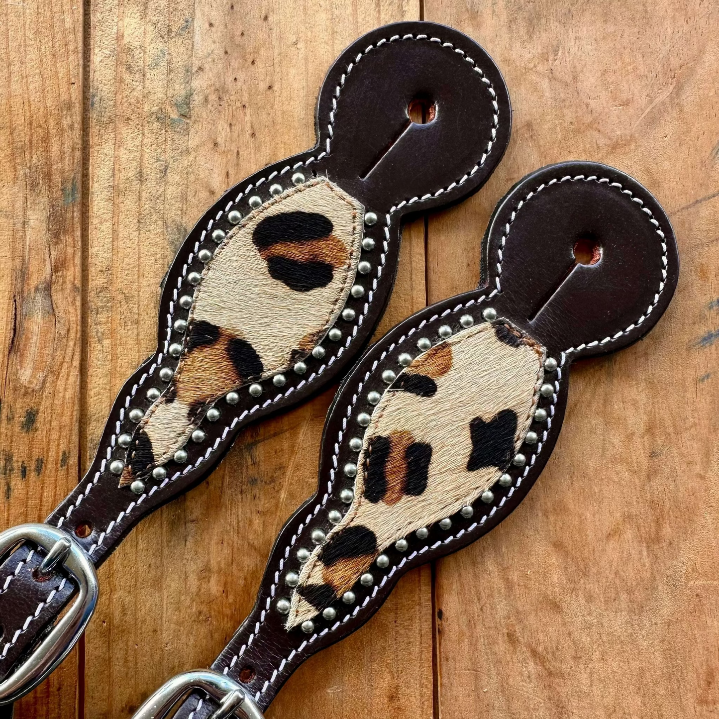 Two leather dog collars with leopard-print hair-on-hide panels and silver-tone studs, displayed on a rustic wooden background.