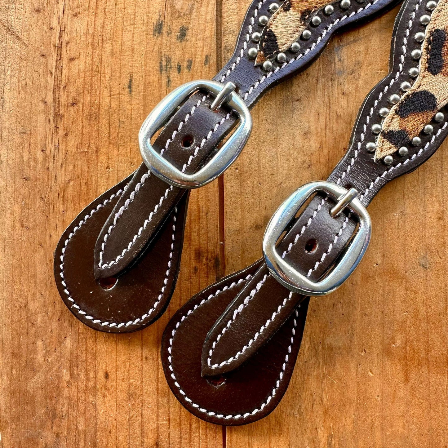 A close-up of a leather strap accessory laid flat on a wooden surface, featuring dark brown leather with white stitching, silver-tone metal buckles, and a leopard-print section with silver studs.