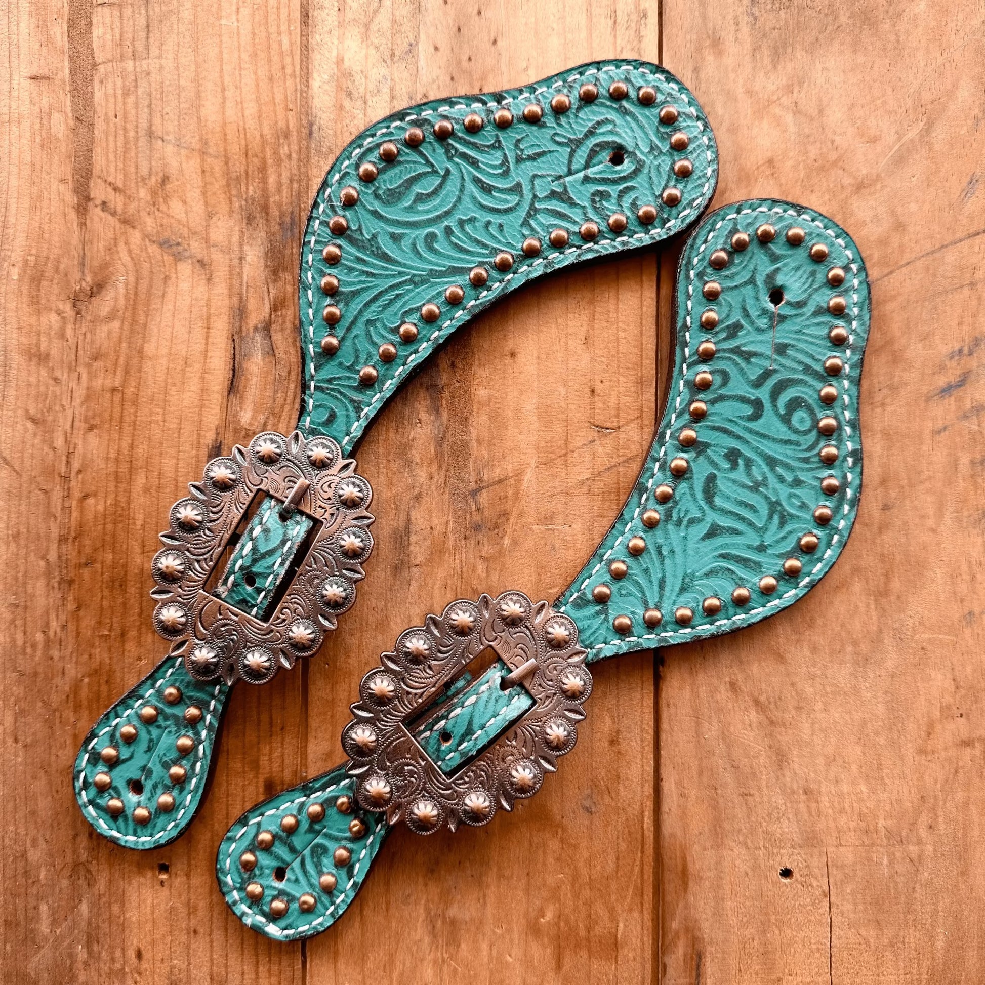 A pair of turquoise tooled leather western-style straps with bronze-colored buckles and copper studs, displayed on a rustic wooden surface.