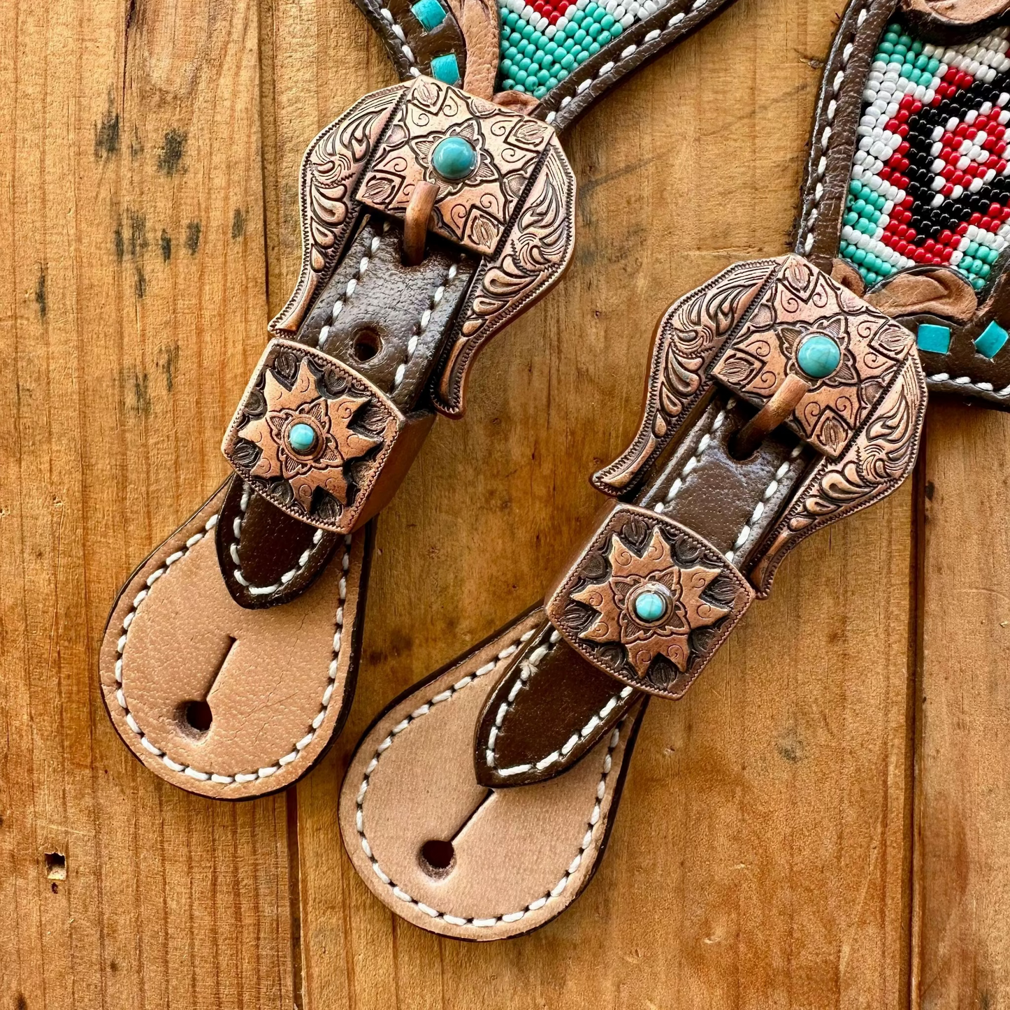 A pair of western-style leather thong sandals with hand-tooled brown leather straps, turquoise stone accents, and a multicolor beaded strap detail, displayed on a rustic wooden surface.